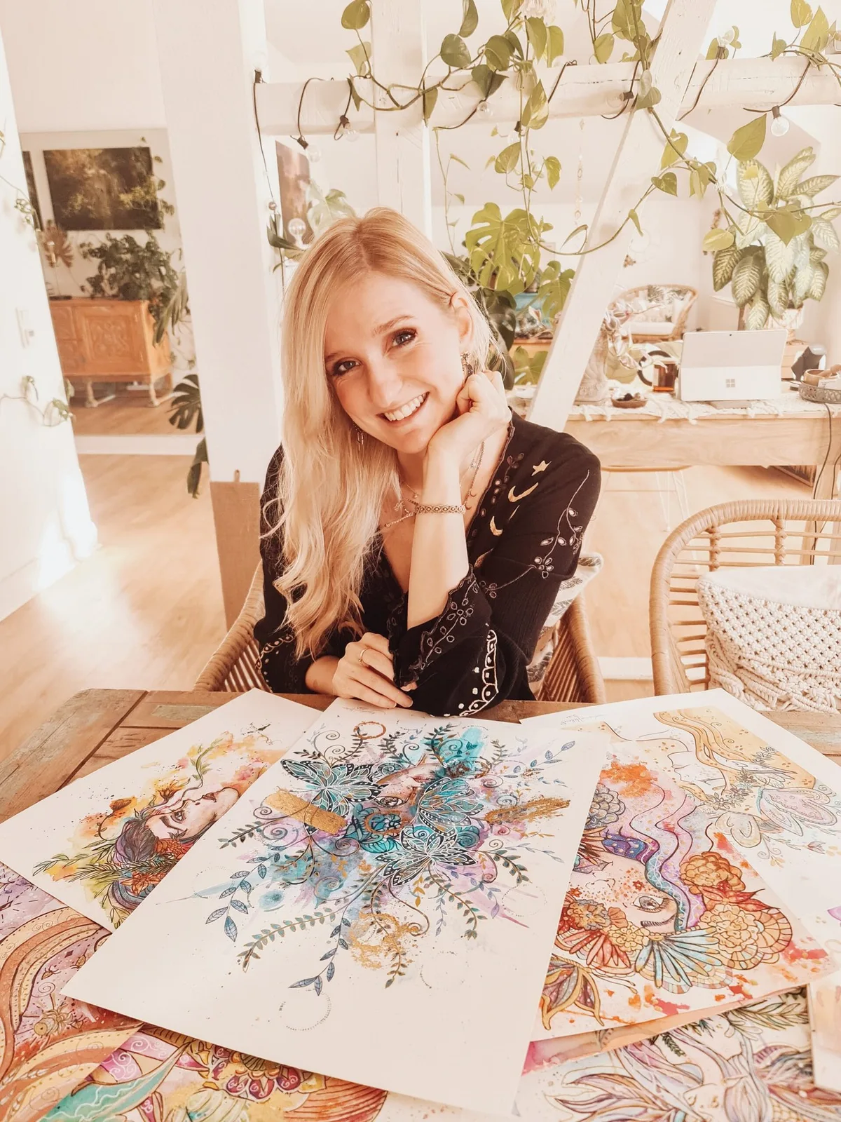 Laura Helena sitzt lächelnd in ihrem Atelier vor mehreren selbst gemalten Aquarellen