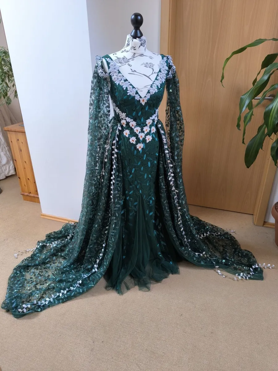 Dunkelgrünes Kleid mit Pailletten-Blättern, weißen Blüten und langen Spitzenärmeln auf einer Schneiderpuppe